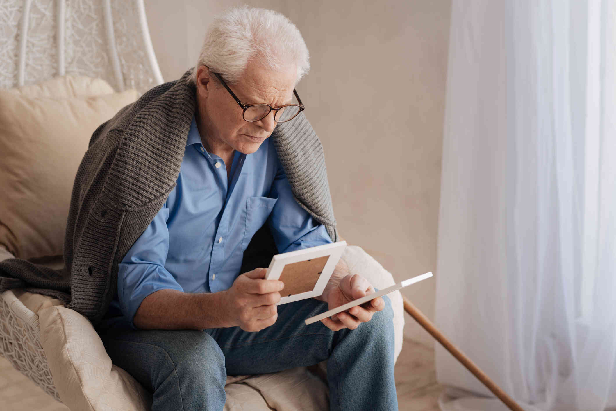 An elderly man seated on his couch, thoughtfully looking at photos he is holding.